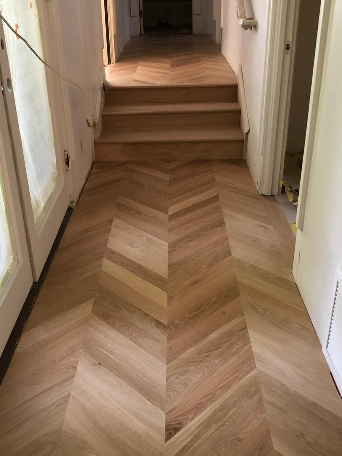 Acacia Wood Floors project — completed warm oak chevron-pattern wood floor in a hallway continuing onto stairs with white walls