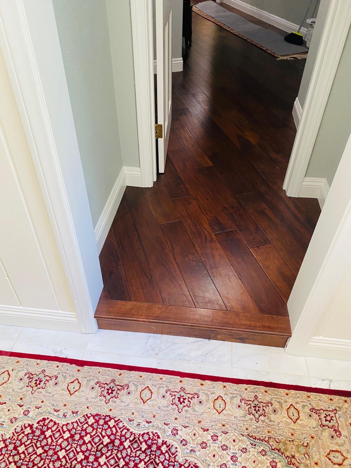 Completed Acacia Wood Floors project — rich dark walnut hardwood floor continuing through a doorway into another room with light marble tile, framed by crisp white trim and a red patterned runner rug in the foreground