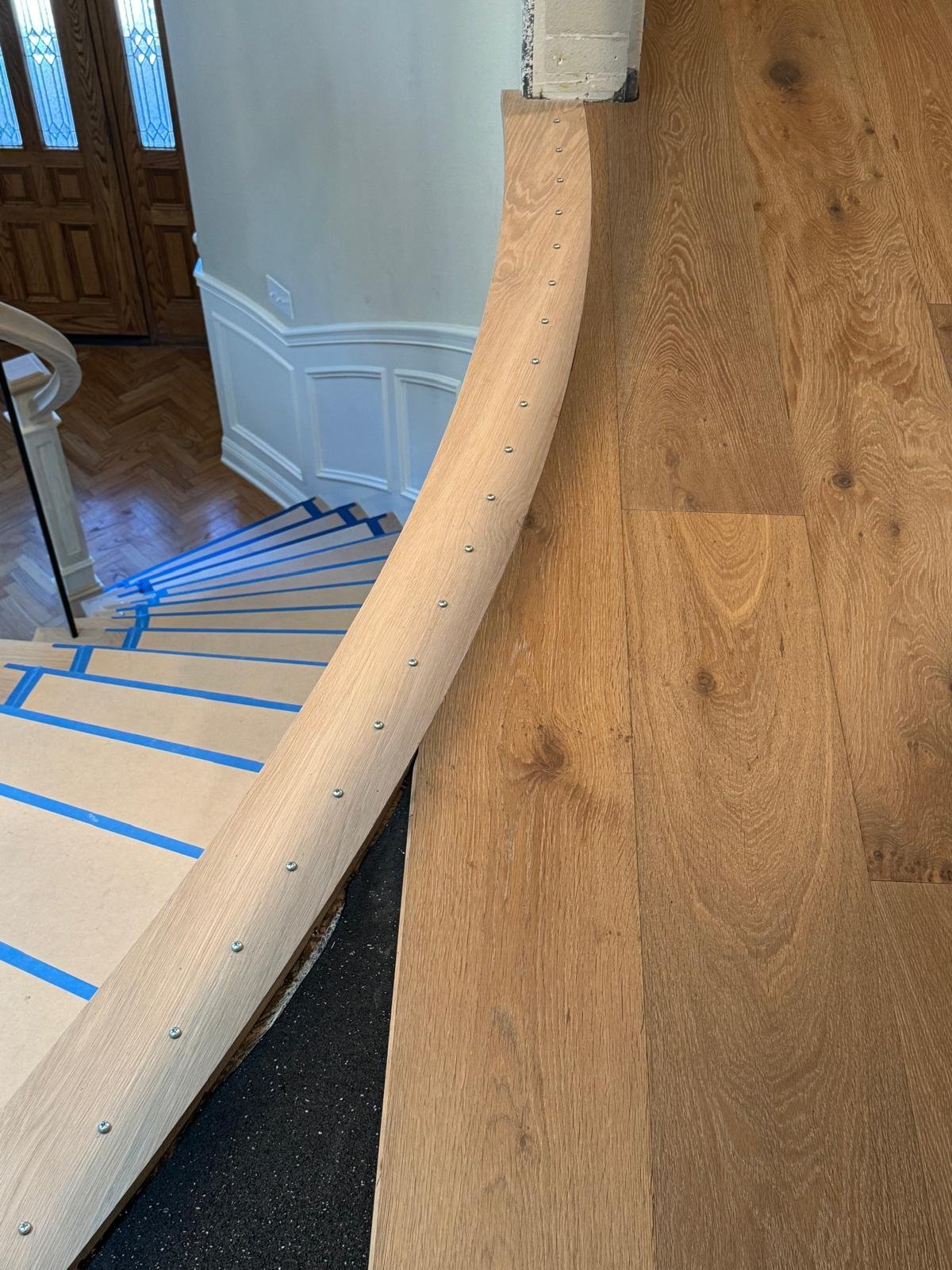 Acacia Wood Floors stair and floor installation in progress — custom light-oak curved handrail with chrome metal fasteners along a grand staircase, freshly installed wide-plank oak floor on the landing, painter's tape protecting the risers, and an elegant leaded-glass entry door and herringbone parquet entryway below