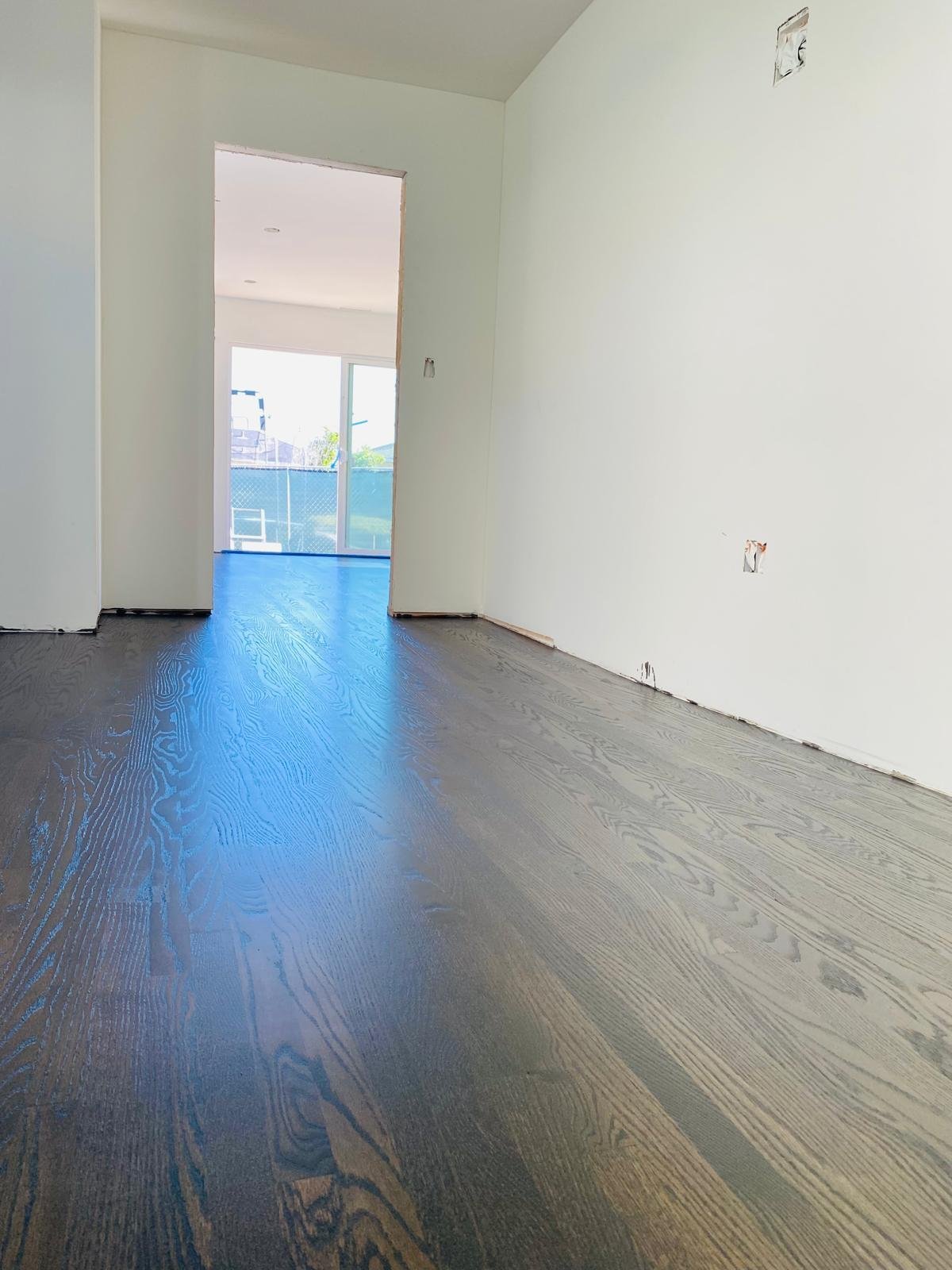 Acacia Wood Floors refinish project in progress — dark-stained wide-plank oak floor in a freshly painted open floor plan, with the deep grain pattern picking up reflections of natural light from the doorway and window, white walls and clean baseboard line