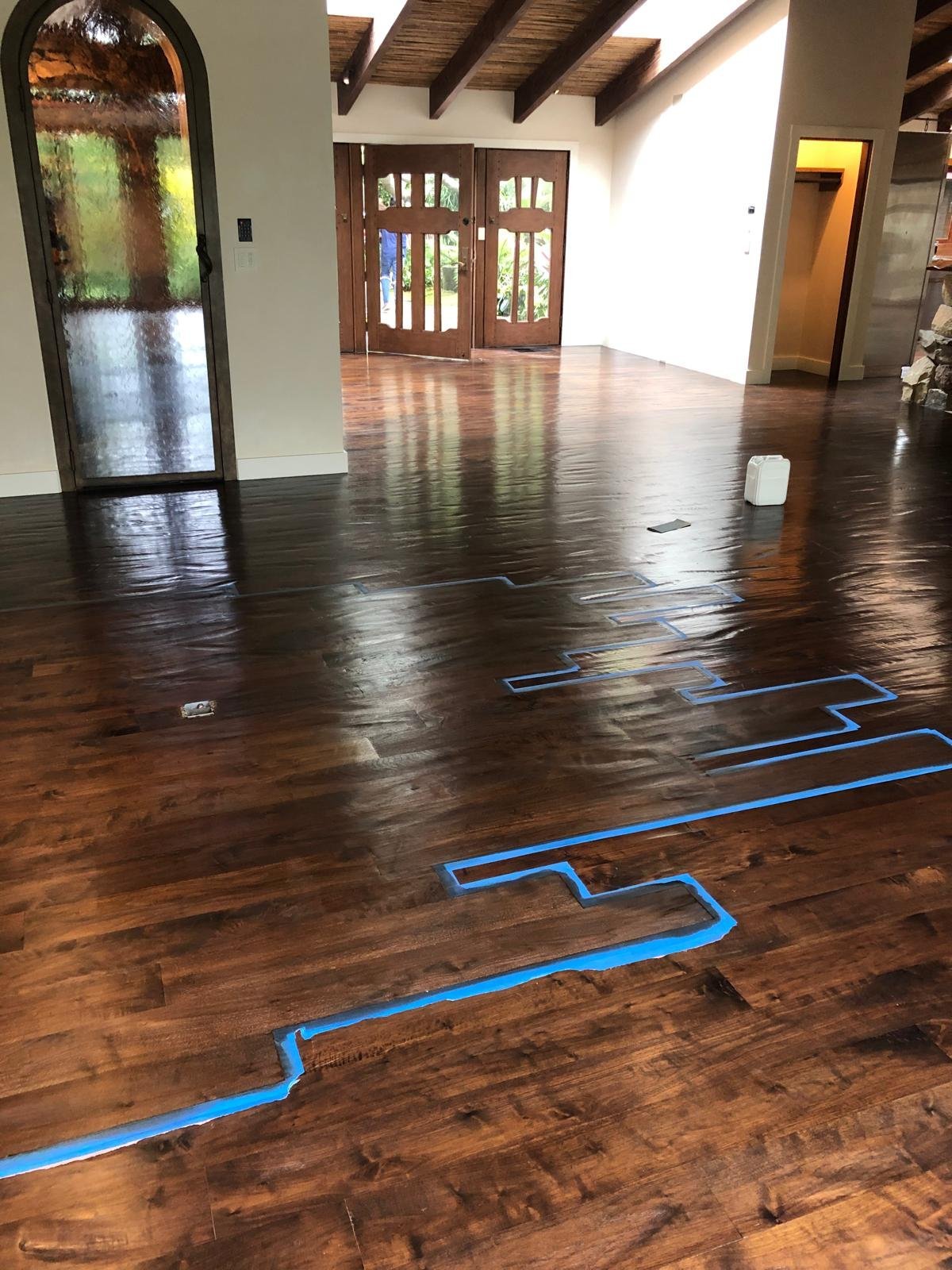 Acacia Wood Floors project in progress — spacious interior with rich dark walnut hardwood floors, blue painter's tape outlining the layout pattern, exposed wood ceiling beams, arched glass door and large windows