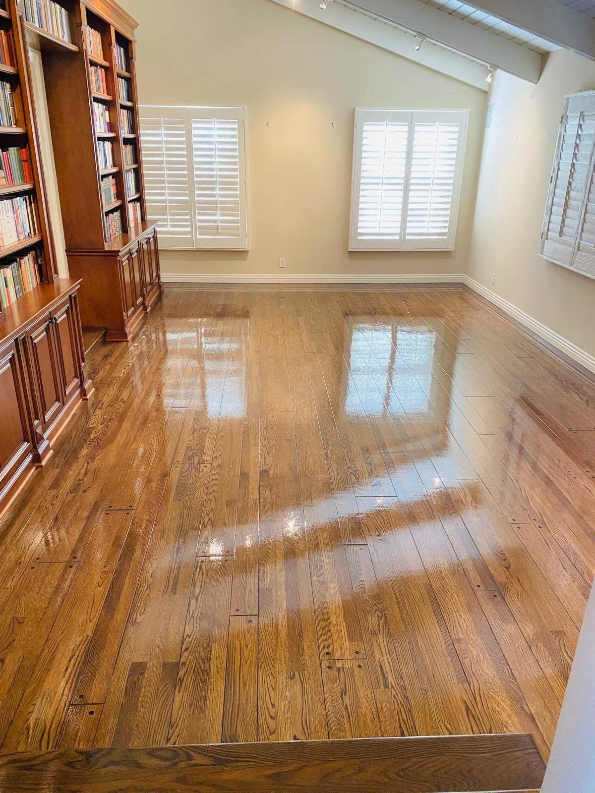 Completed Acacia Wood Floors project — spacious empty room with rich honey-toned oak hardwood floor with high-gloss refinish, custom dark wood built-in bookshelves, and white shuttered windows