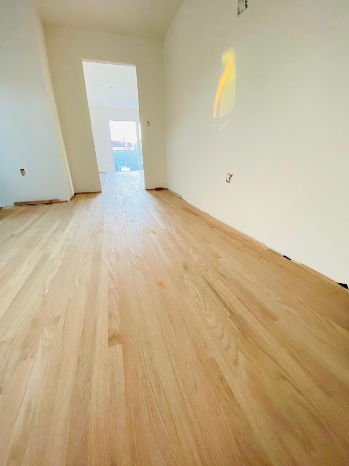 Completed Acacia Wood Floors project — newly installed wide-plank light-oak hardwood floor in a freshly painted room with white walls, open doorway, and natural sunlight streaming in to highlight the rich grain