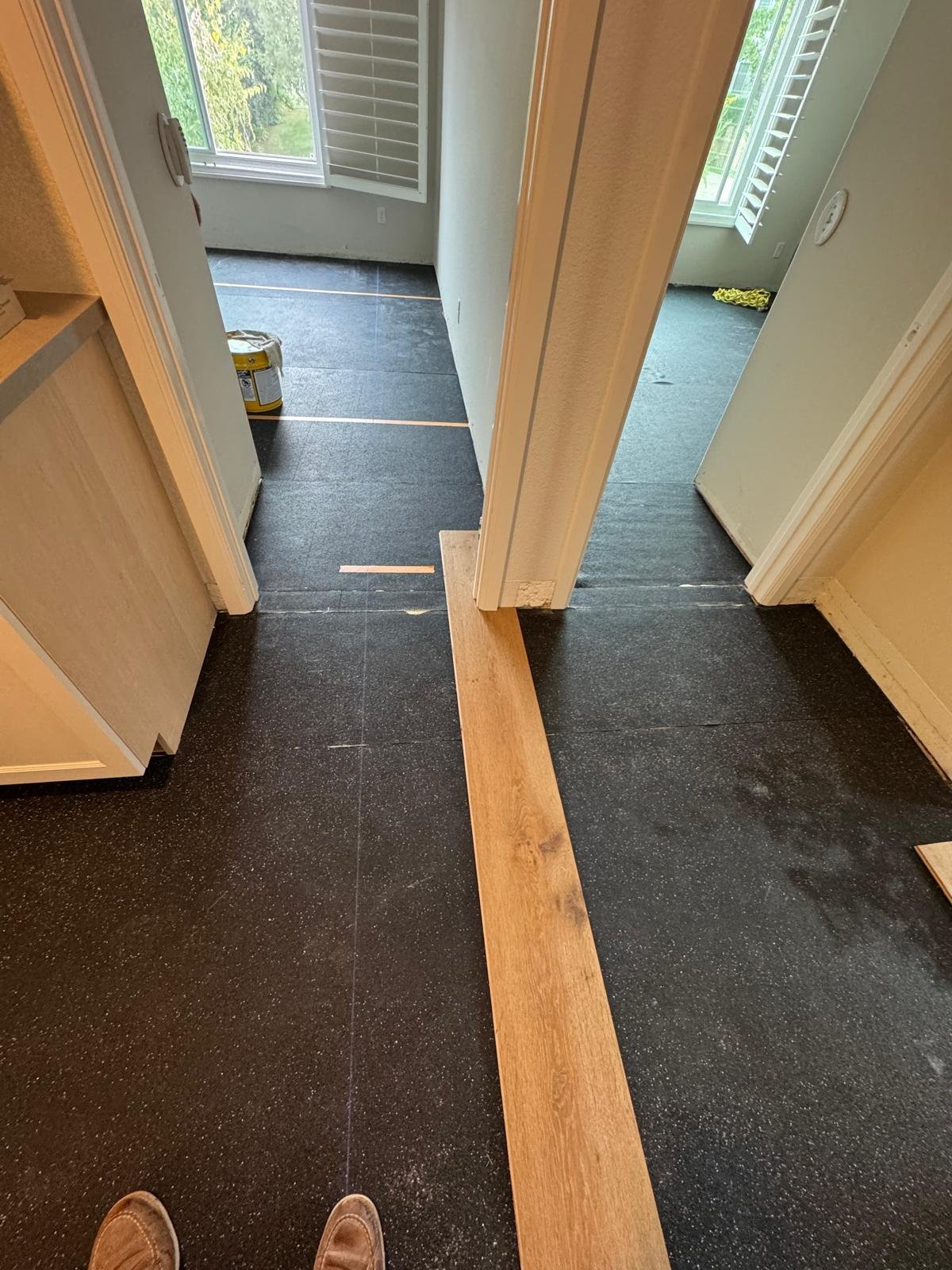 Acacia Wood Floors floor prep project in progress — interior hallway covered with black asphalt-saturated felt underlayment, with a freshly cut light-oak threshold transition strip laid in the doorway between two rooms, an Acacia team member's suede shoes visible in the foreground, paint can and shutters in the background
