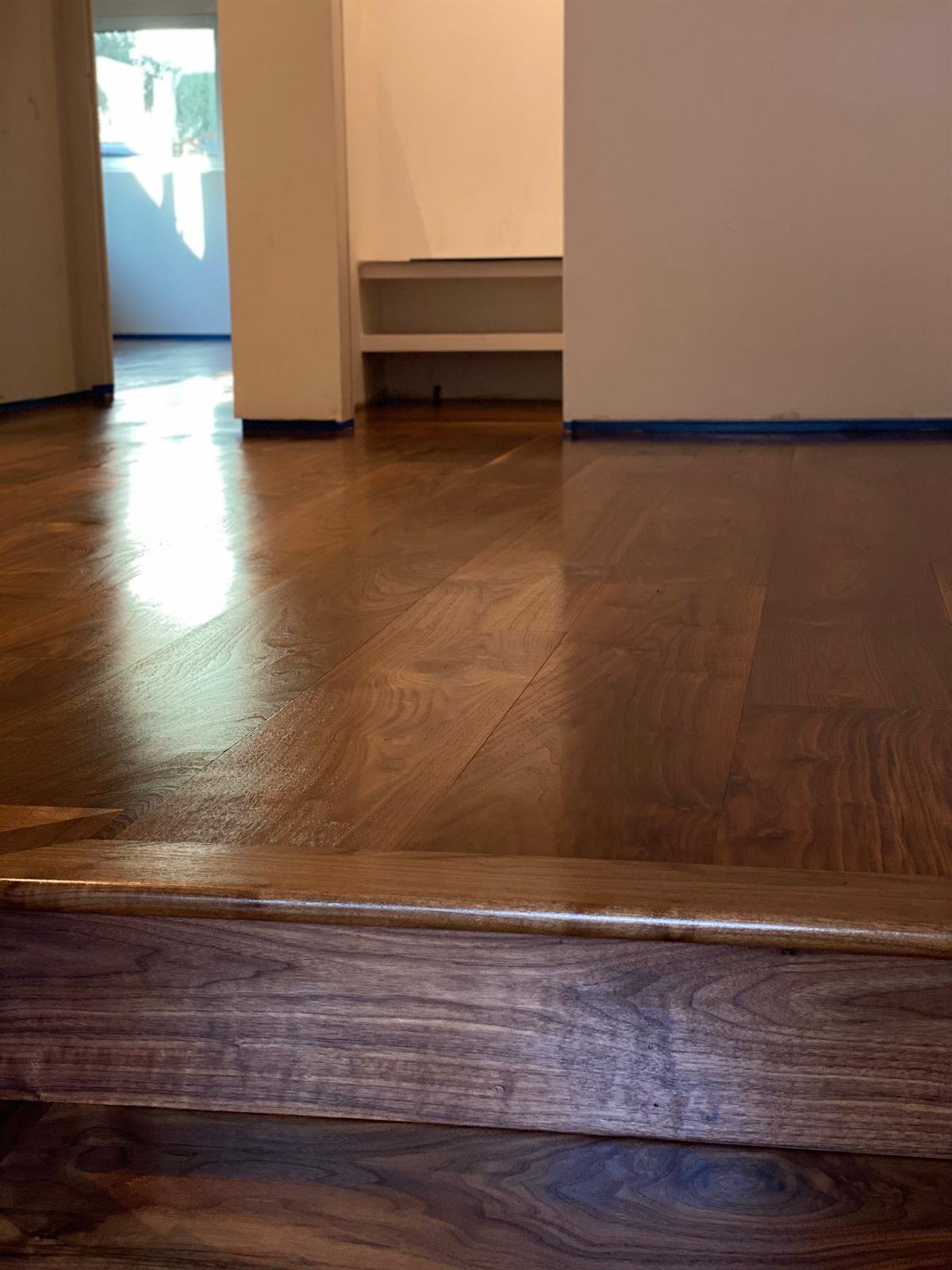 Completed Acacia Wood Floors project — bright hallway with warm honey-toned oak hardwood floor, cream walls, built-in shelving, blue painter's tape along the baseboards, and natural light from the doorway