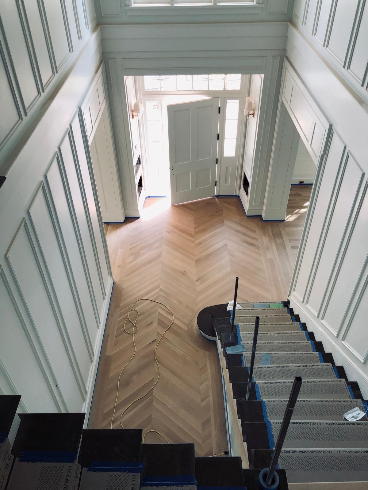 Acacia Wood Floors project in progress — elegant entryway with fresh chevron-pattern light oak hardwood flooring, white wainscoting, staircase with protective step covers leading to open front door