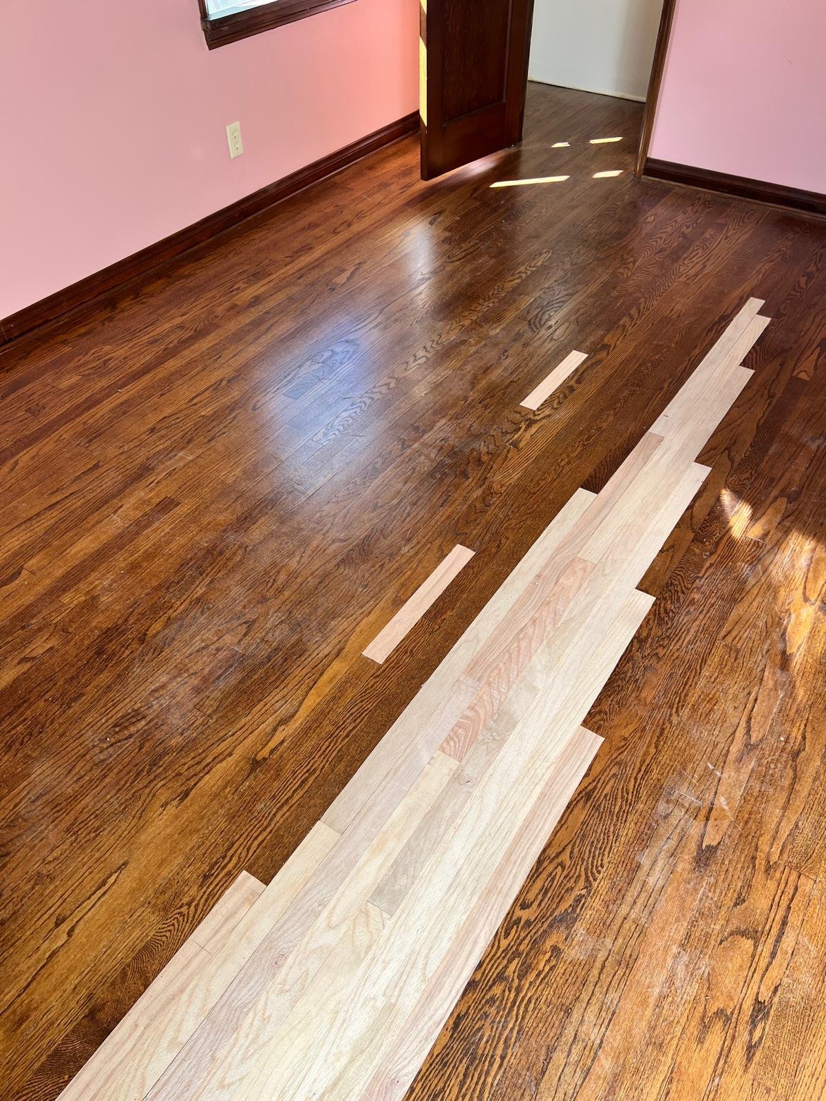 Acacia Wood Floors floor repair in progress — new light-oak replacement planks laid into a dark walnut hardwood floor showing the water-damaged section being restored, with pink walls and burgundy door in the background