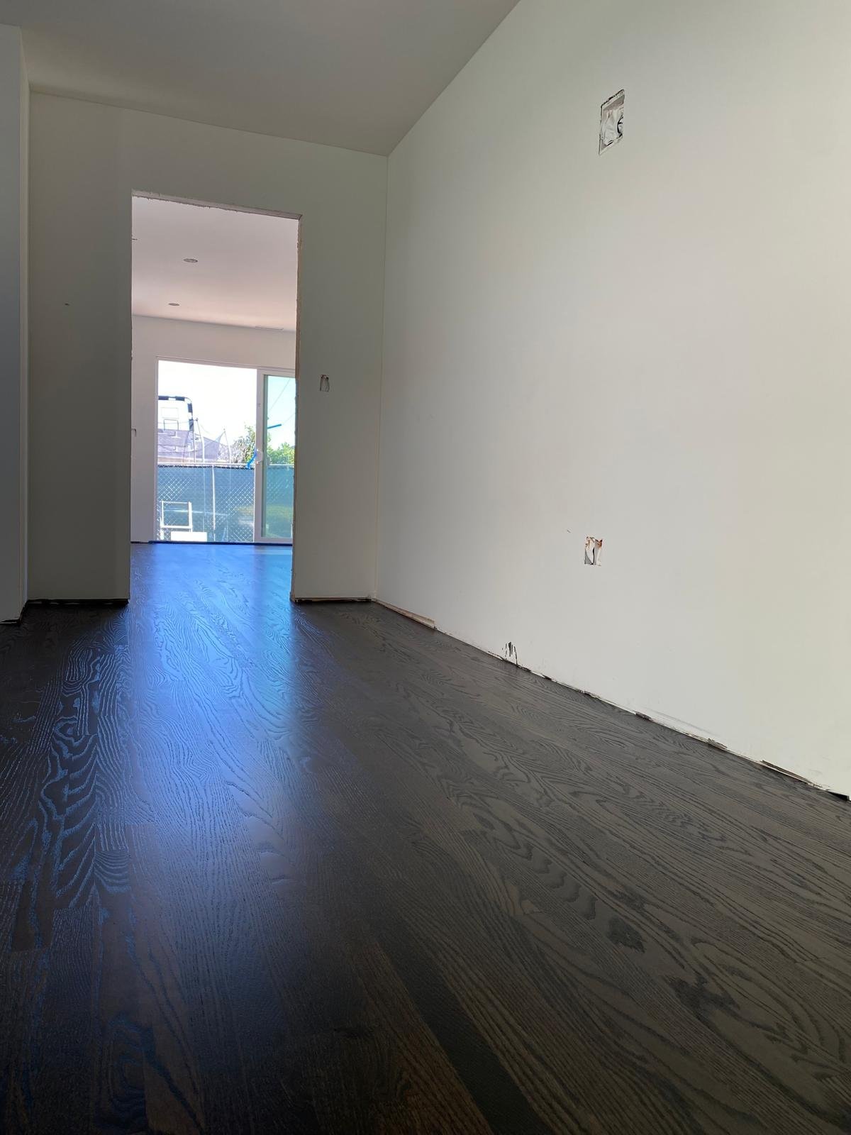 Completed Acacia Wood Floors refinish project — freshly refinished dark-stain wide-plank oak hardwood floor with a high-gloss mirror finish reflecting natural light from a sliding glass door and window, white freshly painted walls, and clean baseboard line in an open empty room