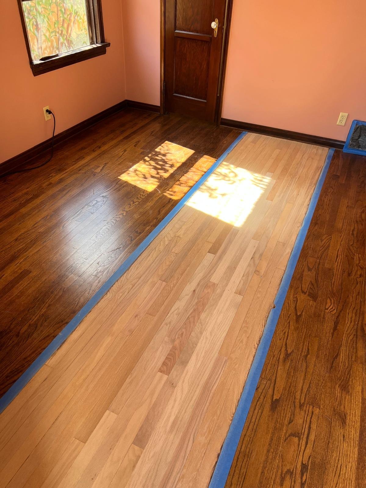 Acacia Wood Floors hardwood refinish in progress — stunning side-by-side comparison of the same floor with blue painter's tape dividing the dark walnut finished section from the fresh light-honey natural oak being refinished, orange walls, dark wood door and natural light