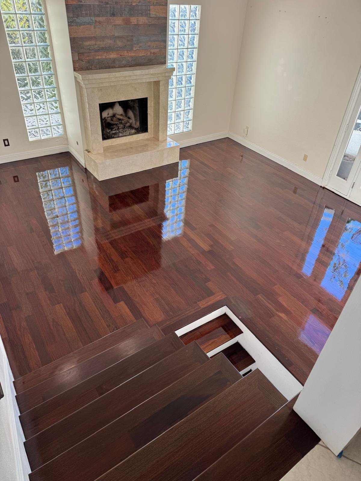 Completed Acacia Wood Floors project — stunning two-story living room with high-gloss deep walnut hardwood floors reflecting blue-tinted glass block windows, cream stone fireplace with reclaimed-wood accent wall, matching walnut staircase with white risers in the foreground
