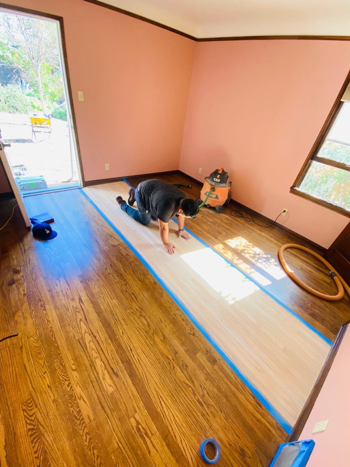 Acacia Wood Floors refinishing project in progress — Acacia team member on hands and knees refinishing a honey-oak hardwood floor with blue painter's tape protecting sections of the wood, professional sander and hose in the salmon-colored room with large windows