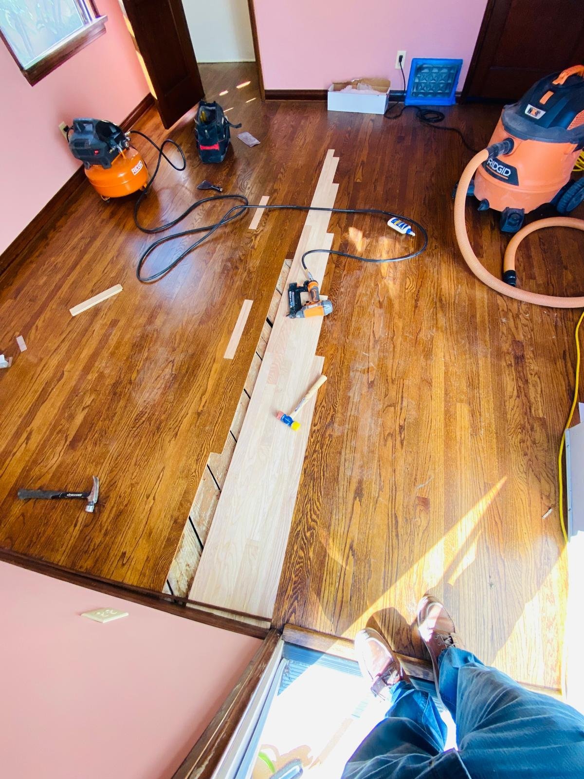 Acacia Wood Floors floor repair in progress — overhead view of new light-oak replacement planks being fitted into a dark walnut floor, with the Acacia team member's boots, tools, and professional equipment in frame