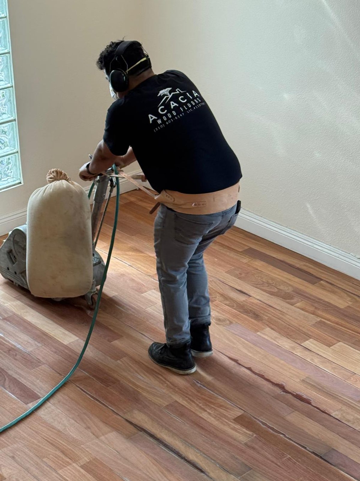 Pedro of Acacia Wood Floors sanding a hardwood floor in person — wearing the Acacia Wood Floors t-shirt with ear protection and a dust mask, operating a professional drum-style floor sander with dust-collection bag on a freshly refinishing red-oak floor in an empty room