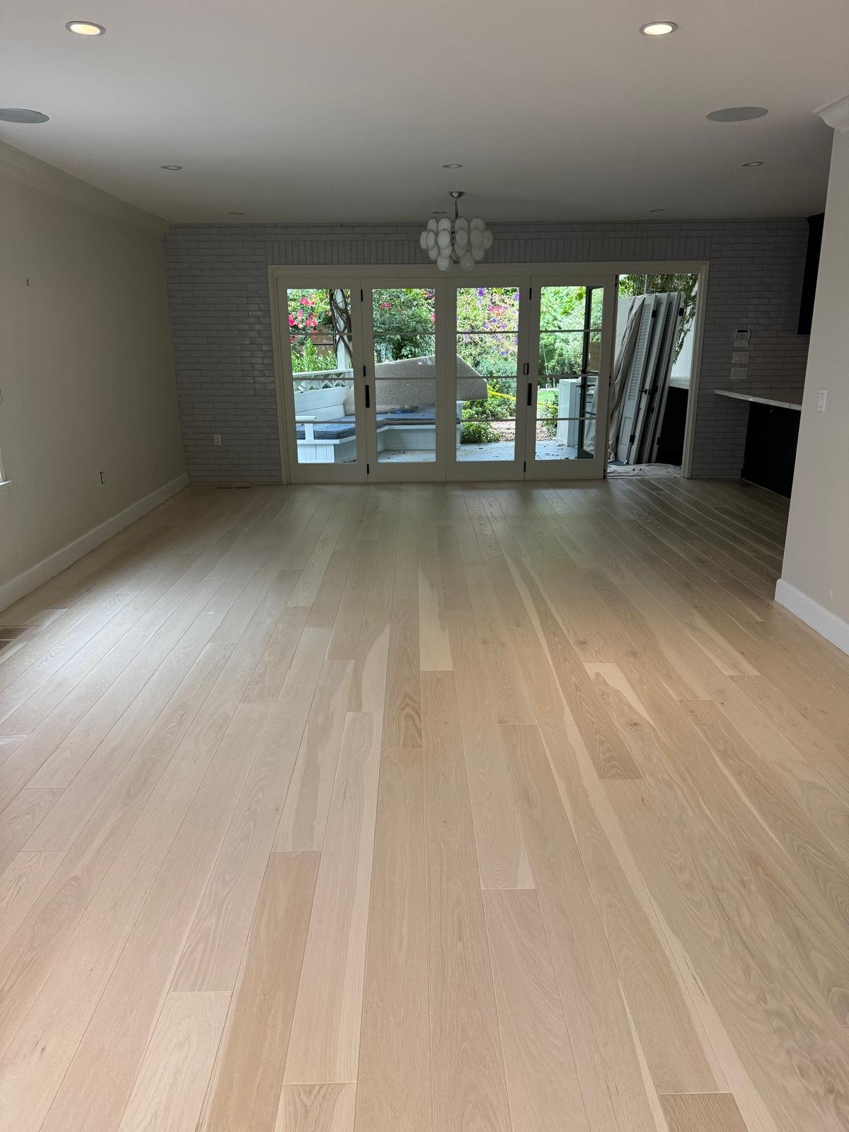 Completed Acacia Wood Floors project — bright empty living room with newly installed wide-plank light-oak hardwood floor, white brick fireplace wall, French glass doors opening to a green garden patio with flowering vines, and a modern chandelier overhead